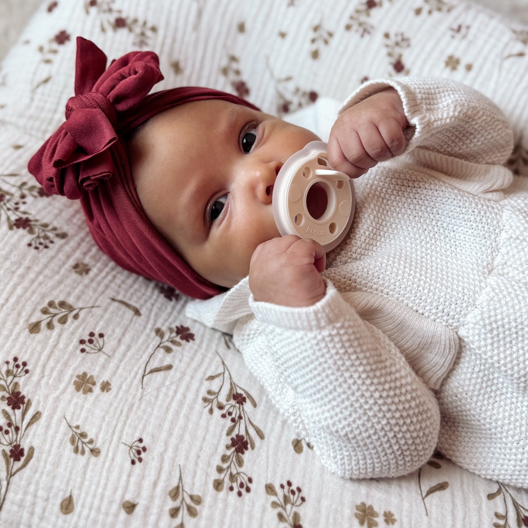 Baby lying on a floral cushion wearing a white outfit and red head wrap with a creamy white, "pearl" mōmi pacifier.