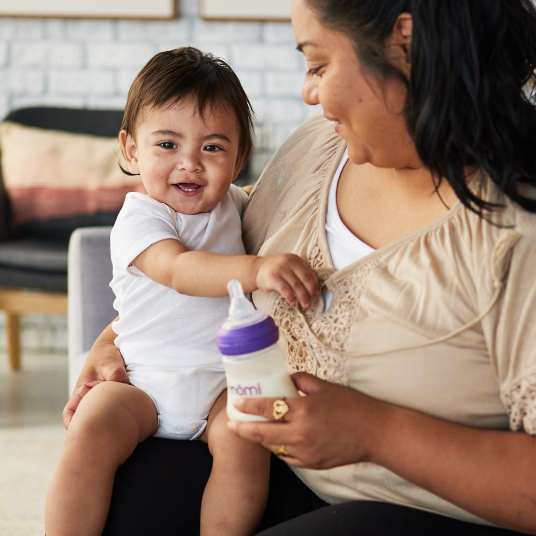 Woman holding a baby and a momi baby bottle in a living room setting