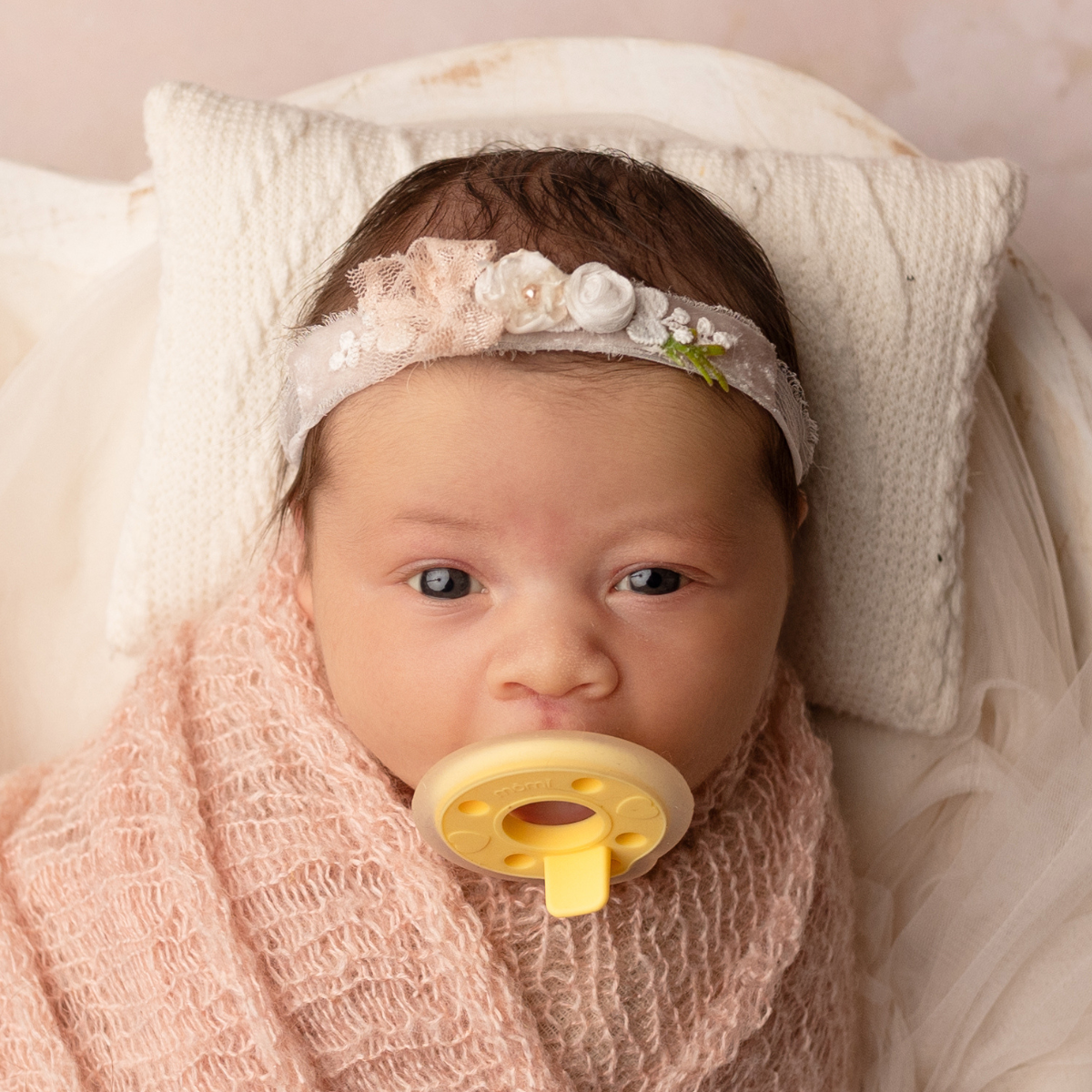 Baby girl wrapped in pink blanket with floral headband and yellow mōmi pacifier, lying on white pillow.